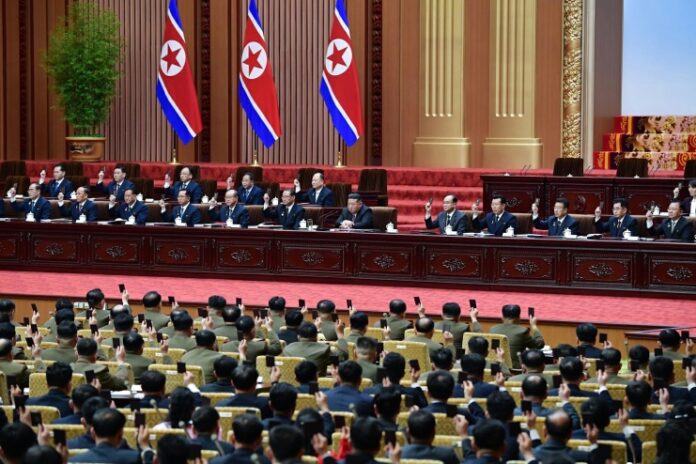 Kim Jong Un and senior North Korean officials raise hands in a vote at the presidium table during the first session of the 15th Supreme People's Assembly in Pyongyang, with rows of delegates and military officers raising hands and phones in the hall below, and three North Korean flags displayed behind the platform.