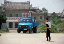 A blue FAW truck parked in front of a building at Chongsan-ri Farm in Kangso, North Korea, with a child walking in the foreground.