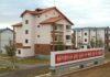 A newly built apartment block with red tile roofing and balconies stands among similar housing in a landscaped development, with a red propaganda sign in the foreground reading a slogan calling to concentrate efforts on socialist economic construction and accelerate the revolution's advance.
