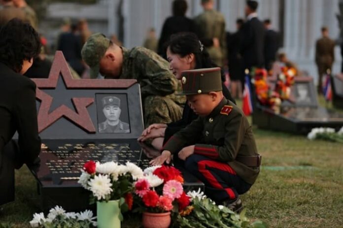 Family members in military and civilian dress kneel and bow at gravestones bearing red stars and photographs of soldiers, with flowers laid at the graves and a memorial building in the background, at North Korea's Overseas Military Operations Combat Merit Memorial Hall.