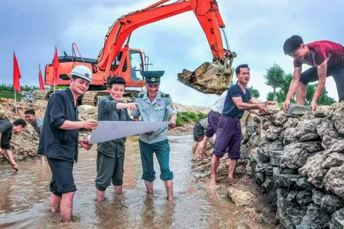 North Korean workers and a uniformed official review plans during river and embankment repair work, with an excavator operating in the background
