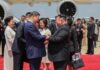 Chinese President Xi Jinping and North Korean leader Kim Jong Un shake hands and clasp arms on a red carpet beside Xi's plane at Pyongyang International Airport, with Peng Liyuan and Ri Sol Ju standing nearby alongside officials and photographers.