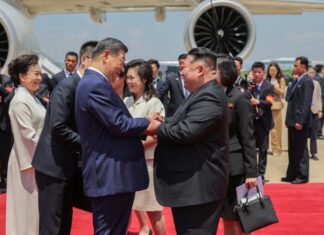 Chinese President Xi Jinping and North Korean leader Kim Jong Un shake hands and clasp arms on a red carpet beside Xi's plane at Pyongyang International Airport, with Peng Liyuan and Ri Sol Ju standing nearby alongside officials and photographers.