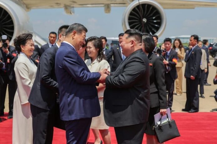 xi-jinping-kim-jong-un-pyongyang-airport-arrival Chinese President Xi Jinping and North Korean leader Kim Jong Un shake hands and clasp arms on a red carpet beside Xi's plane at Pyongyang International Airport, with Peng Liyuan and Ri Sol Ju standing nearby alongside officials and photographers.