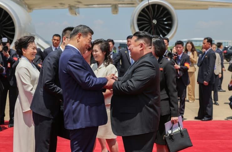 Hyesan residents see hope for economic relief in Xi Jinping’s visit Chinese President Xi Jinping and North Korean leader Kim Jong Un shake hands and clasp arms on a red carpet beside Xi's plane at Pyongyang International Airport, with Peng Liyuan and Ri Sol Ju standing nearby alongside officials and photographers.