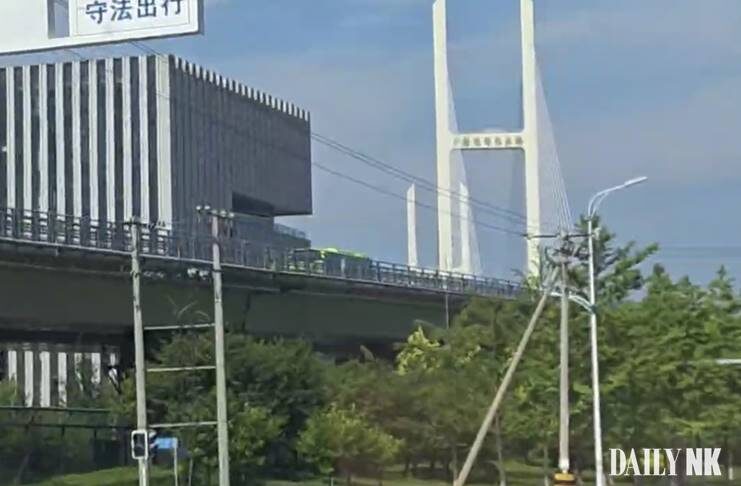 A green bus travels along an elevated approach road leading to the New Yalu River Bridge in Dandong, China, with the bridge's white cable-stayed tower visible in the background.